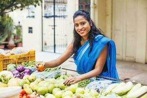 lindo jovem indiano mulher vestindo saree selecionando tomates a partir de uma vegetal impedir dentro uma mercado foto