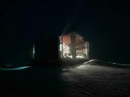 cabine dentro inverno noite. lado luz a partir de a noite janela do a chalé em uma inverno Prado com montes de neve. foto