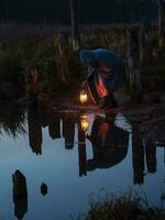 dobrado homem segurando a velho luminária ao ar livre perto a lago. mão detém uma ampla luminária dentro a escuro. antigo querosene lanterna ilumina a caminho em uma noite. luz e esperança conceito. foto