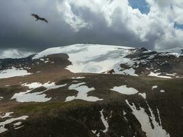 brilhante atmosférico cenário em topo do visto neve montanha cume debaixo Sombrio nuvens. lindo panorama com mosca Águia acima montanha alcance com luz solar debaixo tormentoso denso nuvens. foto