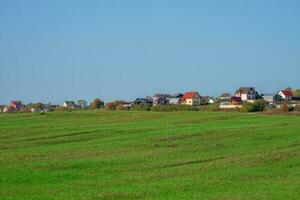 uma verde Primavera campo dentro frente do uma moderno Vila em uma Colina contra uma Claro azul céu. agrícola terra. foto