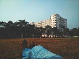 pessoa em jeans azul deitada em um campo de grama marrom olhando para um prédio branco de vários andares