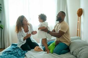 feliz africano americano pais com pequeno filhas jogando em a cama dentro quarto às lar, feliz família conceito foto