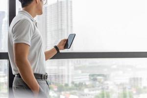 empresário em pé ao lado de uma grande janela em um prédio moderno, procurando um telefone celular