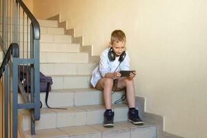 uma feliz estudante é sentado em a escadas com fones de ouvido e olhando às a telefone saltando Aulas foto