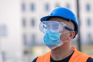 trabalhador da construção civil usando capacete azul, colete reflexivo e máscara cirúrgica protetora foto