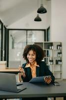 confiante o negócio especialista atraente sorridente jovem mulher digitando computador portátil ang segurando digital tábua em escrivaninha dentro escritório. foto