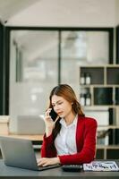 o negócio ásia mulher falando em a telefone e usando uma computador portátil com uma sorrir enquanto sentado às moderno escritório foto
