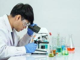 ásia masculino aluna cientista vestindo uma médico vestido dentro a laboratório, olhando às a resultados através uma microscópio. em uma mesa dentro uma científico pesquisa laboratório com teste tubos em uma branco mesa foto