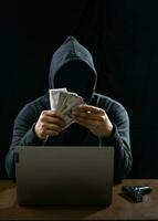 hacker espião homem vestindo uma Preto camisa, sentado em uma cadeira e uma mesa, é uma ladrão, mãos segurando dinheiro, contando a montante obtido a partir de sequestro ou roubando, dentro uma breu sala. foto