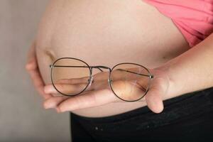 jovem grávida mulher mantém olho óculos dentro frente do dela barriga. foto