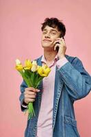 jovem cara dentro uma jeans Jaqueta com uma ramalhete do flores falando em a telefone Rosa fundo foto