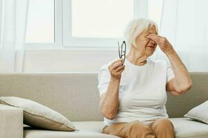 idosos mulher forte olho dor sentado em a sofá, saúde problemas dentro velho idade, pobre qualidade do vida. uma avó com cinzento cabelo detém em para dela dolorido olhos com óculos dentro dela mão. foto