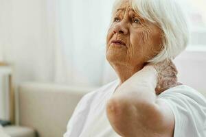 idosos mulher forte pescoço dor sentado em a sofá, saúde problemas dentro velho idade, pobre qualidade do vida. foto