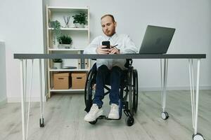 uma homem dentro uma cadeira de rodas uma homem de negocios dentro a escritório trabalho às uma computador portátil e detém uma telefone dentro dele mãos, Consideração, integração para dentro sociedade, a conceito do trabalhando uma pessoa com deficiências foto