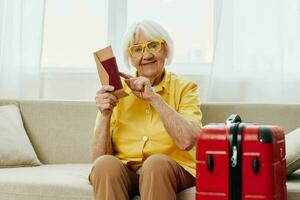 feliz Senior mulher com Passaporte e viagem bilhete embalado uma vermelho mala, período de férias e saúde Cuidado. sorridente velho mulher alegremente sentado em a sofá antes a viagem elevado dela mãos acima dentro alegria. foto