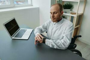 uma homem cadeira de rodas, uma homem de negocios dentro a escritório às uma computador portátil, fadiga e emocional esgotamento às a fim do a trabalhando dia, a conceito do trabalhando uma pessoa com deficiências, liberdade a partir de social frameworks foto