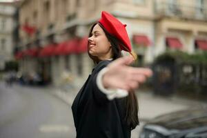 moda mulher sorrir com dentes em pé em a rua dentro frente do a cidade turista Segue mim à moda roupas com vermelho lábios e vermelho boina, viagem, cinematográfico cor, retro vintage estilo, urbano moda. foto