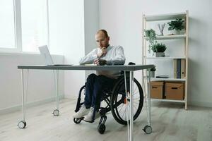 uma homem dentro uma cadeira de rodas, uma homem de negocios dentro a escritório trabalho às uma computador portátil, escrevendo baixa uma plano dentro uma caderno, Consideração, integração para dentro sociedade, a conceito do trabalhando uma pessoa com deficiências foto