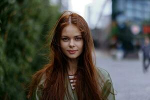 retrato do uma lindo jovem menina dentro a cidade olhando para dentro a Câmera com vermelho vôo cabelo dentro uma verde capa de chuva dentro a cidade contra uma fundo do bambu dentro primavera, estilo de vida dentro a cidade foto