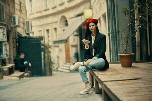 à moda mulher turista dentro à moda roupas dentro uma Jaqueta e vermelho boina sentado lado de fora com telefone dentro mão, célula telefone dentro viagem, francês estilo, cinematográfico cor. foto