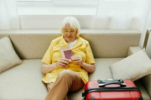 feliz Senior mulher com Passaporte e viagem bilhete embalado uma vermelho mala, período de férias e saúde Cuidado. sorridente velho mulher alegremente senta em a sofá antes a viagem. foto