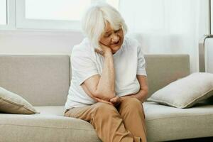 idosos mulher forte pescoço dor sentado em a sofá, saúde problemas dentro velho idade, pobre qualidade do vida. foto