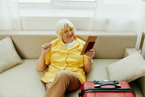 feliz Senior mulher com Passaporte e viagem bilhete embalado uma vermelho mala, período de férias e saúde Cuidado. sorridente velho mulher alegremente senta em a sofá antes a viagem. foto