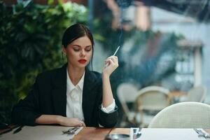 à moda moda mulher retrato sentado dentro uma cafeteria às uma mesa e fumar uma cigarro liberando fumaça a partir de dela boca com vermelho batom, mau hábito, sorrir com dentes e pensativo olhar, cinematográfico vintage foto