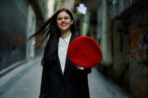 moda mulher sorrir com dentes e Diversão turista dentro à moda roupas dentro Jaqueta e vermelho boina caminhando baixa limitar cidade rua vôo cabelo, viagem, francês estilo, cinematográfico cor, retro vintage estilo. foto