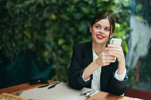 moda mulher blogueiro turista senta dentro uma cafeteria com uma telefone dentro dela mãos lê uma mensagem, Móvel comunicação e a Internet em uma Primavera viagem, vídeo chamar, freelance trabalhos on-line, sorriso, comunicação foto