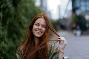 retrato do uma lindo jovem mulher dentro a cidade olhando para dentro a Câmera sorrir com dentes com vermelho vôo cabelo dentro elegante roupas dentro a cidade contra do bambu dentro primavera, estilo de vida dentro a cidade foto