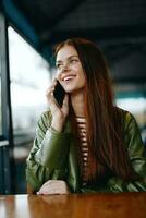 mulher com vermelho cabelo freelance blogueiro sentado dentro uma cafeteria falando em a telefone e sorridente, hipster menina dentro na moda roupas estilo de vida foto