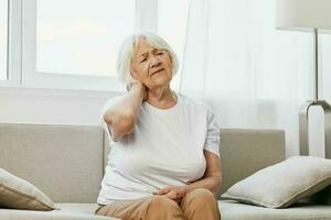 idosos mulher forte pescoço dor sentado em a sofá, saúde problemas dentro velho idade, pobre qualidade do vida. foto