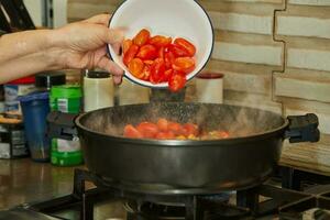 chefe de cozinha lança tomates para dentro uma fritar panela em uma gás fogão foto