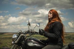 a garota motoqueira ruiva está sentada em um campo de motocicleta em um prado e nuvens foto