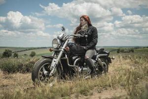 a garota motoqueira ruiva está sentada em um campo de motocicleta em um prado e nuvens