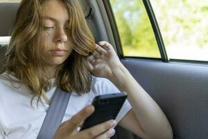 adolescente menina é equitação dentro a costas assento do uma carro e usa Smartphone foto