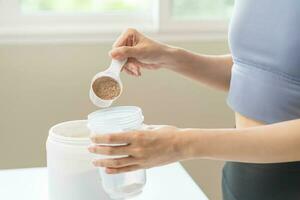 dieta refeição substituição para peso perda, sorrir ásia jovem mulher, menina dentro roupa de esporte, mão dentro segurando colher fazer proteína sacudir, beber suplemento para músculo depois de exercite-se às lar. saudável corpo Cuidado. foto