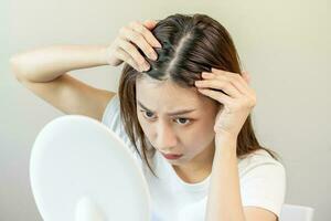 estragado cabelo, face sério ásia jovem mulher preocupação olhando às couro cabeludo dentro espelho, mão dentro pausa para dentro frente cabelo perda, fino problema sintoma às lar. saúde Cuidado xampu beleza, isolado em fundo. foto