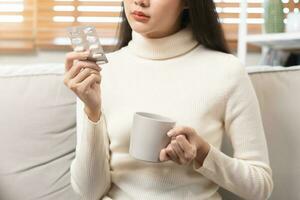 fechar acima febre, doente ásia jovem mulher, menina mão levando medicamento, comprimido para dor de cabeça, estômago dor ou levar vitaminas segurando copo do quente água sentado em sofá às lar. assistência médica, coronavírus pandemia. foto