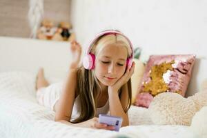 lindo Adolescência menina é relaxante dentro cama usando uma tela sensível ao toque Smartphone, ouvindo para música com uma sem fio fone de ouvido, surfar a Internet dentro a quarto, relaxante confortavelmente, sorridente foto