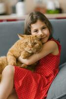 criança menina detém dela animal vermelho britânico gato dentro dela braços e senta com ele em a sofá às casa foto