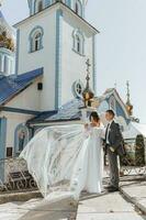 Casamento dia. noivo e noiva segurando mãos e caminhando perto a Igreja depois de a Casamento cerimônia. uma casal do recém-casados dentro amar. debaixo a Maravilhoso claro. gotas a véu foto