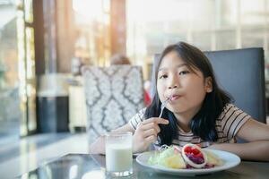 ásia fofa pequeno menina comendo fresco tomate e salada em manhã. foto
