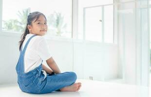 fofa menina vestindo jeans macacão sentado dentro a branco casa, foto