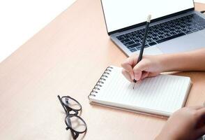 aluna escrevendo dever de casa em em branco caderno com computador portátil em mesa, foto