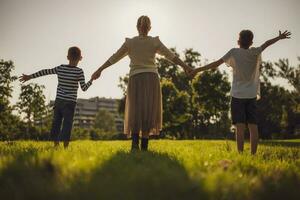feliz mãe é jogando com dela filhos dentro parque. elas estão segurando mãos e desfrutando pôr do sol. foto
