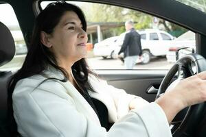 lado retrato do maduro o negócio mulher dentro elegante roupas, sentado dentro moderno carro dentro motorista assento, sorridente olhando atentamente enquanto dirigindo. negócios, bem sucedido pessoas, transporte conceito. foto