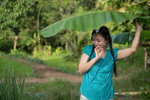 imagem borrada retrato de uma jovem asiática com cabelo preto segurando uma folha de bananeira na chuva no fundo do jardim verde foto